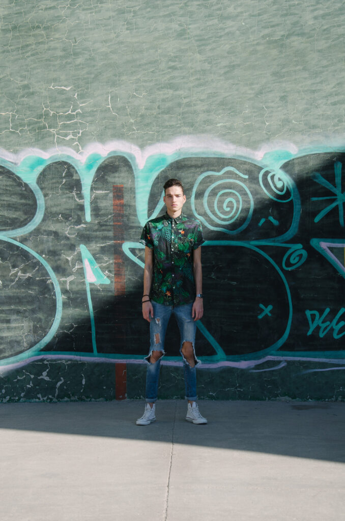 Victor Zepeda stands confidently against a graffiti-covered wall wearing a vibrant tropical shirt and distressed jeans with a watch on his wrist.